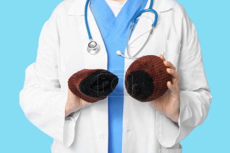 Mature female doctor with stethoscope and knitted model of breast on blue background, closeup. Breast feeding concept