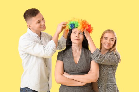 Foto de Gente de negocios poniendo peluca de payaso en su colega contra el fondo amarillo. Celebración del Día de los Inocentes - Imagen libre de derechos