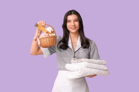 Happy female chambermaid with Easter basket and stack of towels on purple background