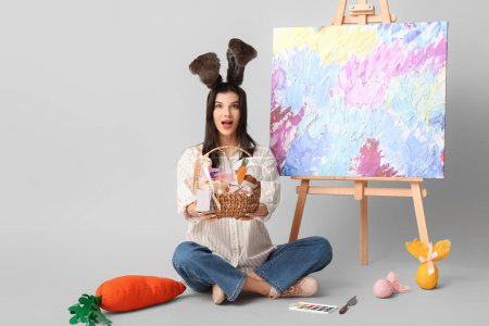 Schockierte Künstlerin in Hasenohren mit Osterkorb, Schokoladeneiern und Staffelei auf grauem Hintergrund