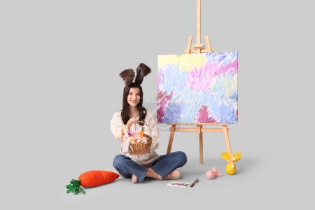 Glückliche Künstlerin in Hasenohren mit Osterkorb, Schokoladeneiern und Staffelei auf grauem Hintergrund