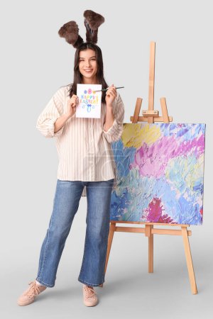 Glückliche Künstlerin in Hasenohren mit Ostergrußkarte, Pinsel und Staffelei auf grauem Hintergrund
