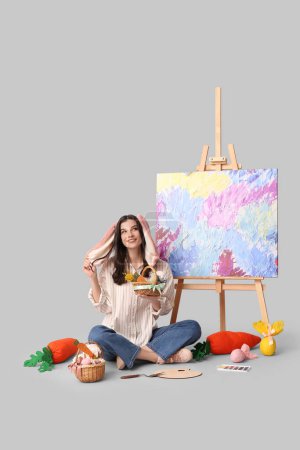 Glückliche Künstlerin in Hasenohren mit Osterkörben, Spielzeugmöhren und Staffelei auf grauem Hintergrund