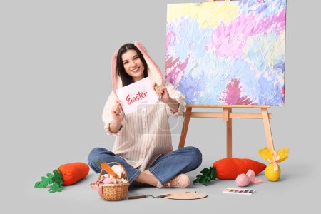 Glückliche Künstlerin in Hasenohren mit Osterkorb, Grußkarte und Staffelei auf grauem Hintergrund