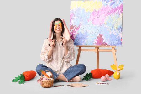 Glückliche Künstlerin in Hasenohren mit Osterkorb, Karotten und Staffelei auf grauem Hintergrund