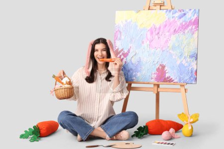 Glückliche Künstlerin in Hasenohren mit Osterkorb, frischer Karotte und Staffelei auf grauem Hintergrund
