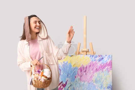 Glückliche Künstlerin in Hasenohren mit Osterkorb, Pinsel und Staffelei auf grauem Hintergrund