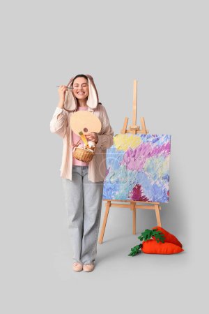 Feliz artista femenina en orejas de conejo con cesta de Pascua, zanahorias de juguete y caballete sobre fondo gris