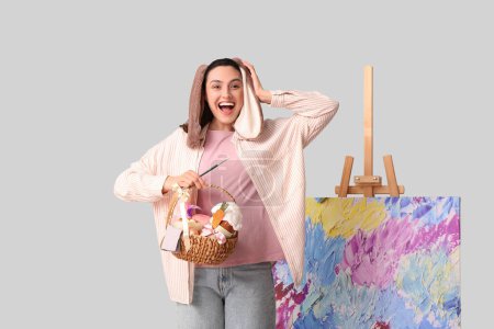 Glückliche Künstlerin in Hasenohren mit Osterkorb, Pinsel und Staffelei auf grauem Hintergrund