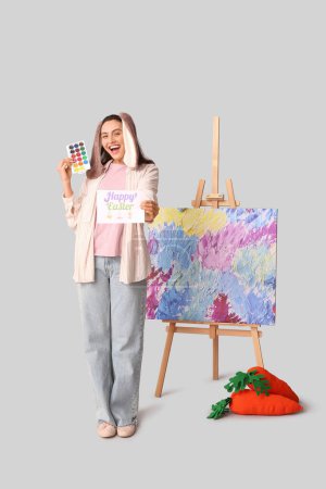 Glückliche Künstlerin in Hasenohren mit Ostergrußkarte, Spielzeugkarotten und Staffelei auf grauem Hintergrund
