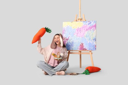 Glückliche Künstlerin in Hasenohren mit Osterkorb, Spielzeugmöhren und Staffelei auf grauem Hintergrund