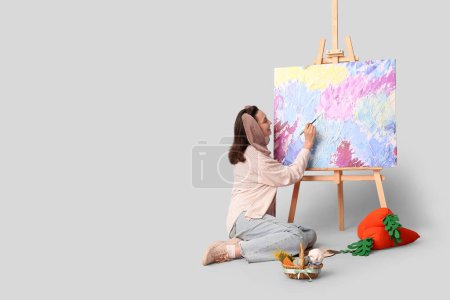 Glückliche Künstlerin in Hasenohren mit Osterkorb, Spielzeugkarotten Malbild auf grauem Hintergrund