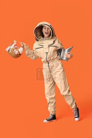 Happy female beekeeper in protective suit with Easter basket and smoker on orange background