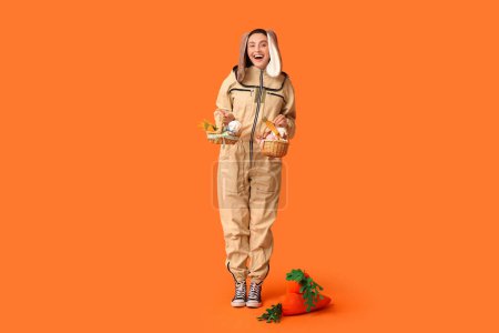 Happy female beekeeper in bunny ears with Easter baskets and toy carrots on orange background