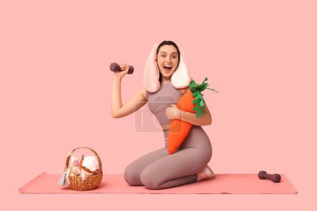 Entrenador de fitness femenino feliz en orejas de conejo con cesta de Pascua, zanahoria de juguete y pesas sobre fondo rosa