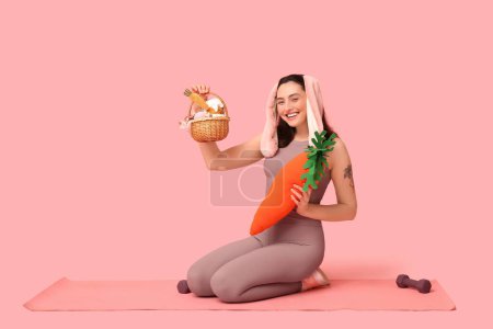 Entrenador de fitness femenino feliz en orejas de conejo con cesta de Pascua, zanahoria de juguete y pesas sobre fondo rosa