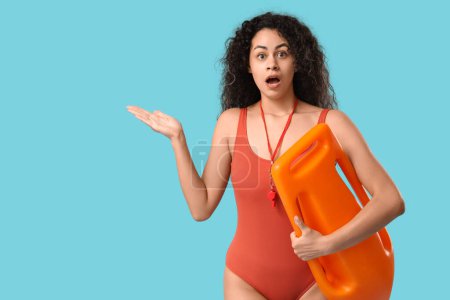 Beautiful young shocked African-American female lifeguard with rescue buoy pointing at something on blue background