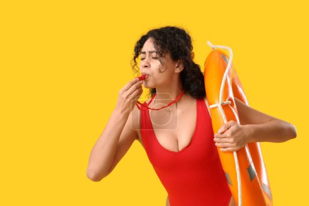 Beautiful young African-American female lifeguard with ring buoy whistling on yellow background