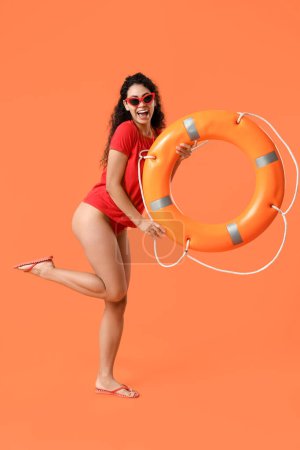 Beautiful young happy African-American female lifeguard with ring buoy on orange background