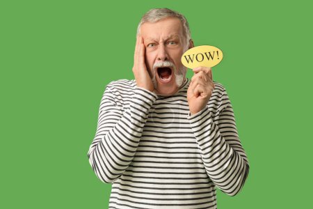 Shocked senior man holding speech bubble with word WOW on green background