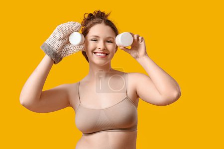 Schöne junge glückliche Frau mit Badehandschuhen und Cremegläsern auf gelbem Hintergrund