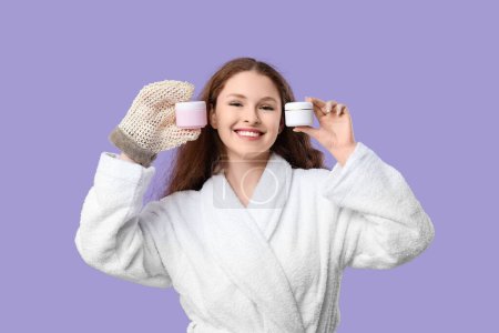 Schöne junge glückliche Frau im Bademantel mit Badehandschuhen und Cremegläsern auf lila Hintergrund