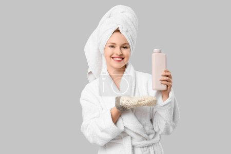 Schöne junge glückliche Frau im Bademantel mit Badehandschuh und Flasche Duschgel auf grauem Hintergrund