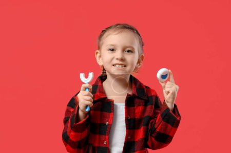 Téléchargez les photos : Joyeux petite fille en pyjama à carreaux avec brosse à dents et fil dentaire sur fond rouge - en image libre de droit