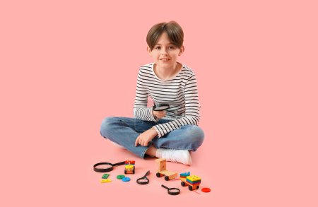 Kleiner Junge mit Lupen und Spielzeug auf rosa Hintergrund
