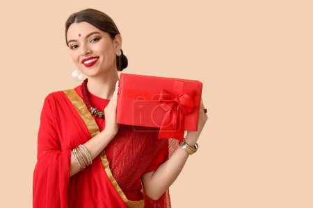 Schöne junge Inderin im Sari mit Geschenkbox auf beigem Hintergrund