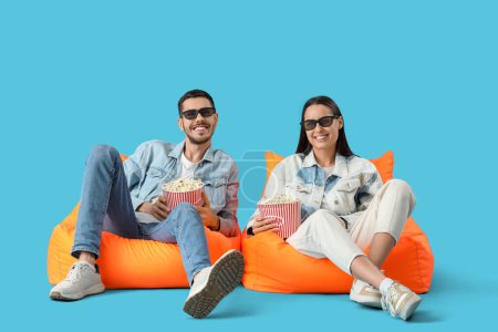 Schönes junges glückliches Paar mit Eimern Popcorn vor dem Fernseher auf Sitzsäcken vor blauem Hintergrund