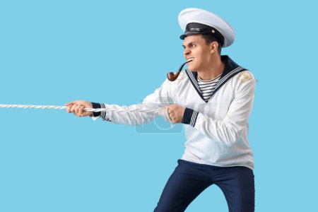 Angry young sailor with smoking pipe pulling rope on blue background