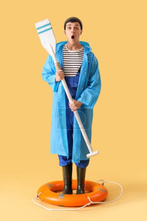 Photo for Shocked young sailor in raincoat with paddle and ring buoy on yellow background - Royalty Free Image