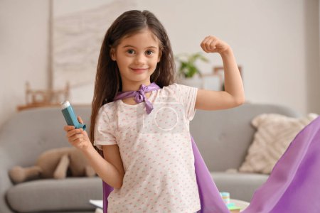 Téléchargez les photos : Petite fille avec cape de super héros et inhalateur montrant les muscles à la maison - en image libre de droit
