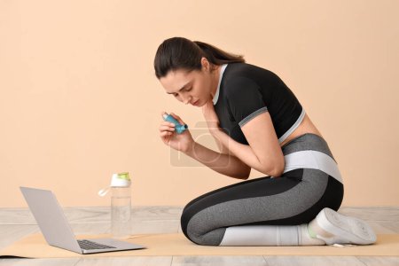 Sporty young woman with inhaler having asthma attack near beige wall