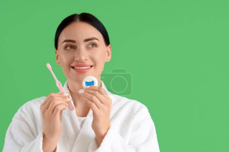 Téléchargez les photos : Belle jeune femme heureuse en peignoir avec fil dentaire et brosse à dents sur fond vert - en image libre de droit