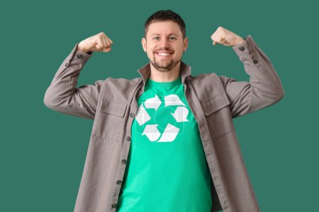 Glücklicher junger Mann im T-Shirt mit Recycling-Logo, das Muskeln auf grünem Hintergrund zeigt. Ökologisches Konzept