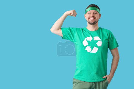 Glücklicher junger Mann im T-Shirt mit Recycling-Logo, das Muskeln auf blauem Hintergrund zeigt. Ökologisches Konzept