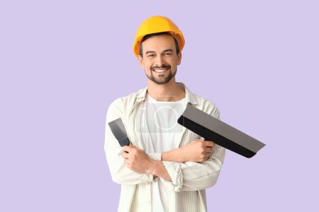 Male decorator with putty knives on lilac background