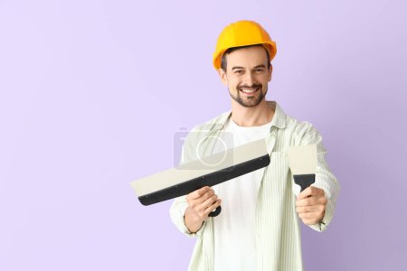 Male decorator with putty knives on lilac background