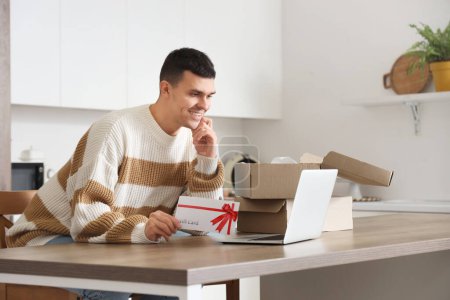 Young man with gift card and laptop shopping online in kitchen