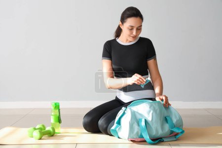 Mujer joven deportiva con inhalador y bolsa sentada cerca de la pared de luz