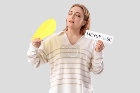 Mature woman holding paper with word MENOPAUSE and fan on light background