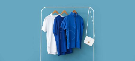 Photo for Stylish t-shirts and bag hanging on rack against blue wall - Royalty Free Image
