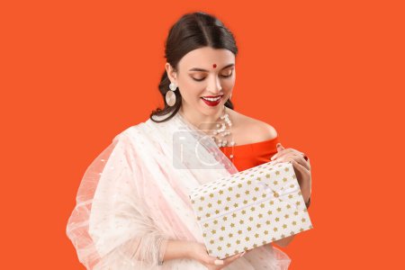 Téléchargez les photos : Belle jeune femme indienne en sari avec boîte cadeau sur fond orange - en image libre de droit