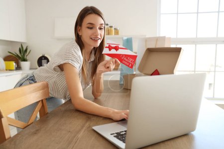 Young woman with gift voucher and laptop shopping online in kitchen