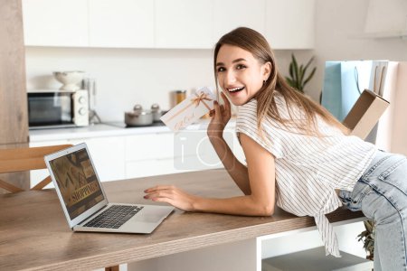 Young woman with gift card and laptop shopping online in kitchen