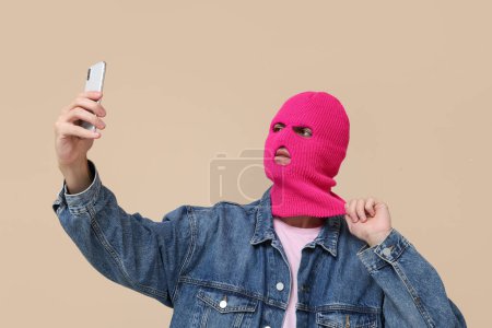 Handsome young man in balaclava taking selfie on beige background
