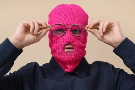 Handsome young man in balaclava with stylish eyeglasses on beige background