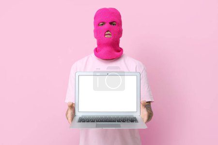 Schöner junger Mann mit Sturmhaube und Laptop auf rosa Hintergrund
